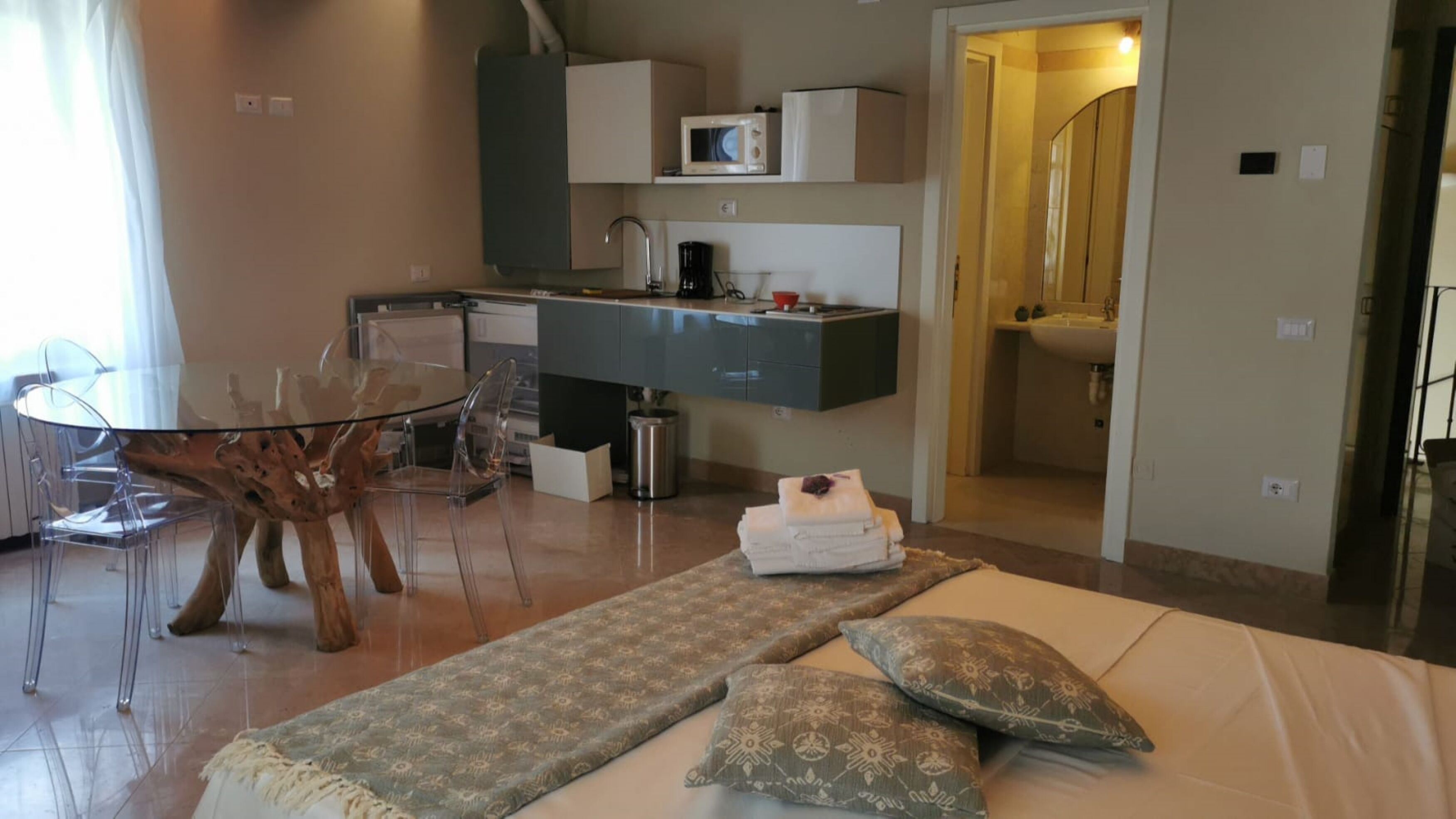A compact hotel room with a kitchenette, a dining area with a glass table and chairs, a bed with a patterned bedspread and pillows, and a bathroom visible through an open door.