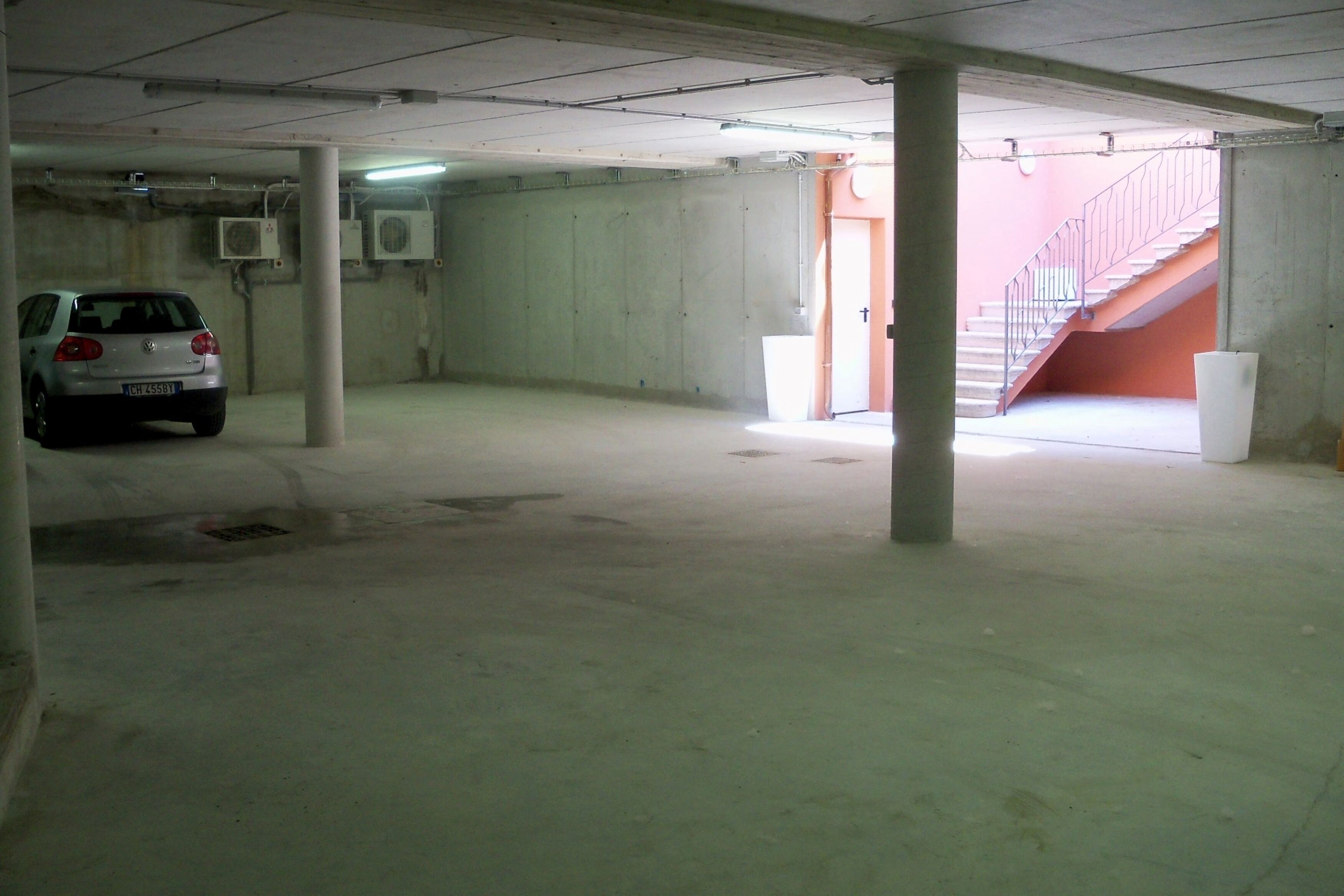 A parking garage with concrete pillars, a white car, and a staircase leading to an upper level.