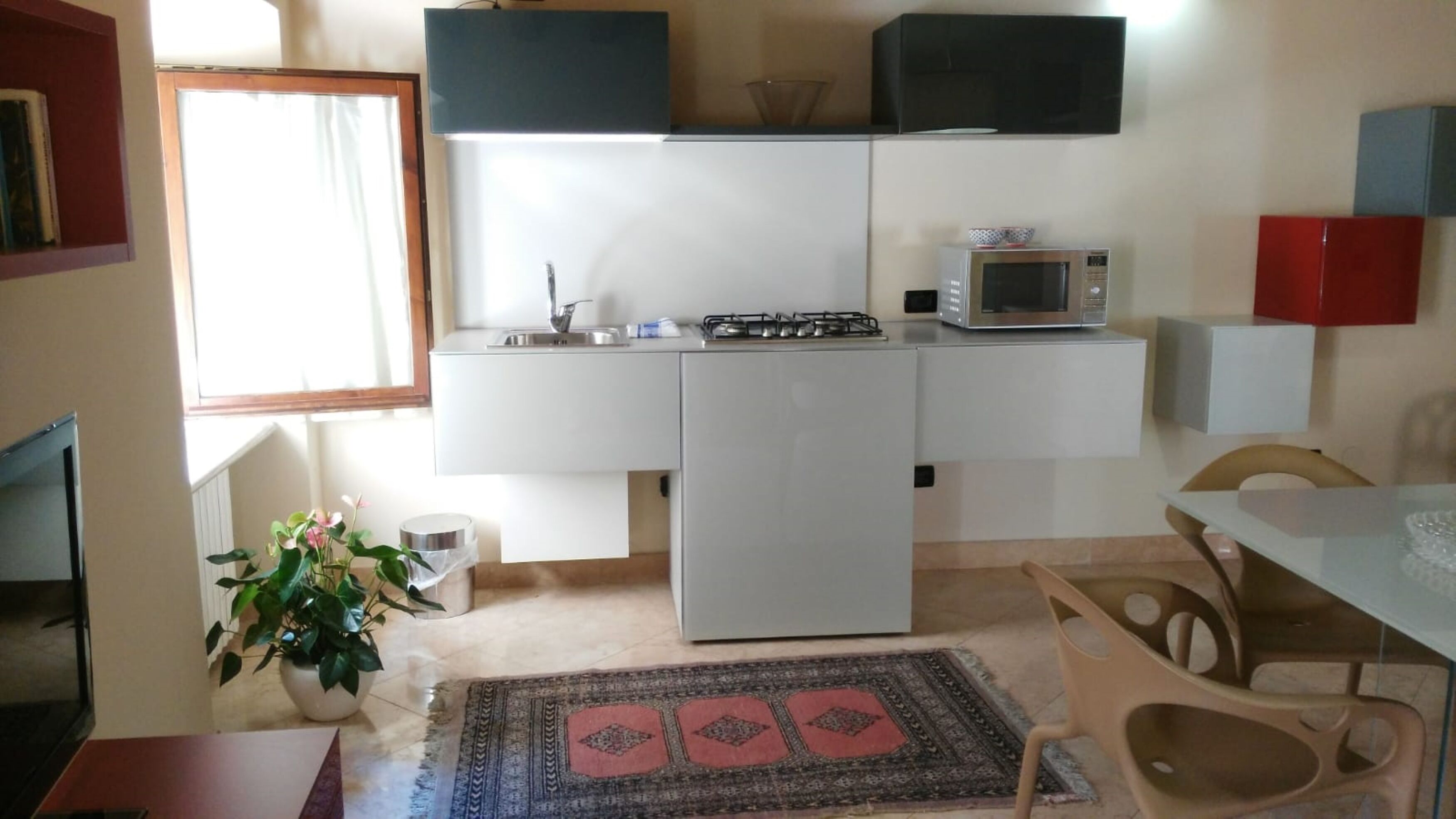 Cocina compacta con estufa a gas, microondas y armarios blancos. Se observa una pequeña mesa de comedor con sillas, una planta en maceta y un tapete estampado.