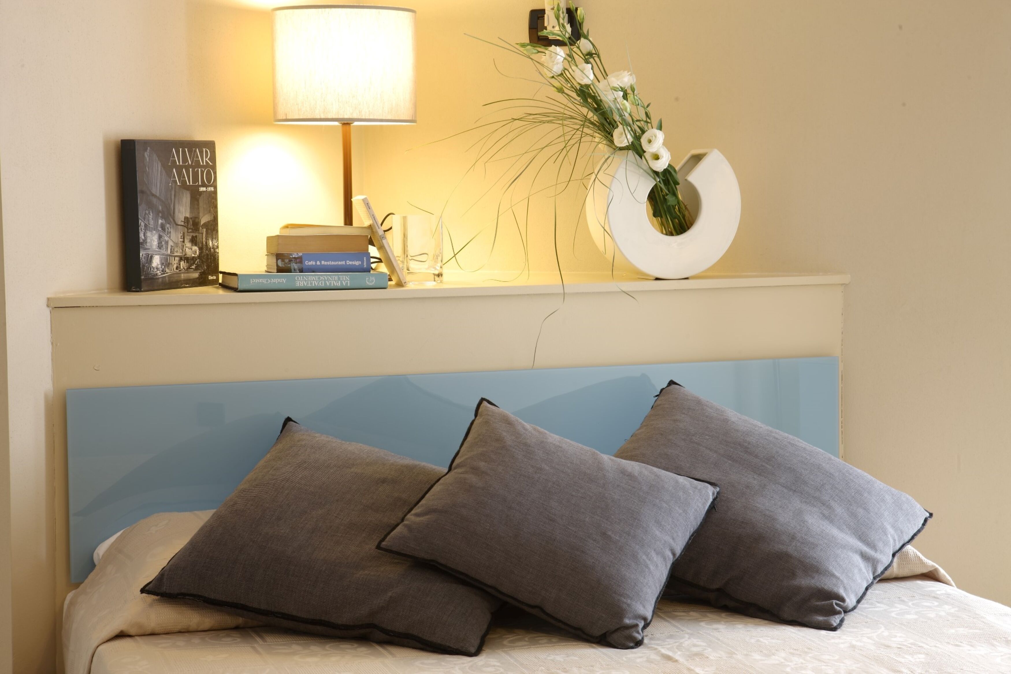 A neatly made bed with two pillows, a bedside table with a lamp, a book, and a decorative vase with flowers.