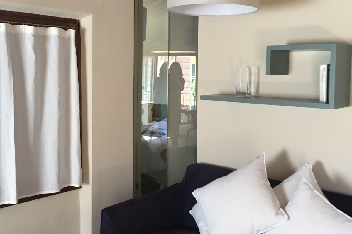 Een kamer met een donkerblauwe bank, witte kussens, een witte hanglamp, een glazen deur en een klein schap met decoratieve objecten.