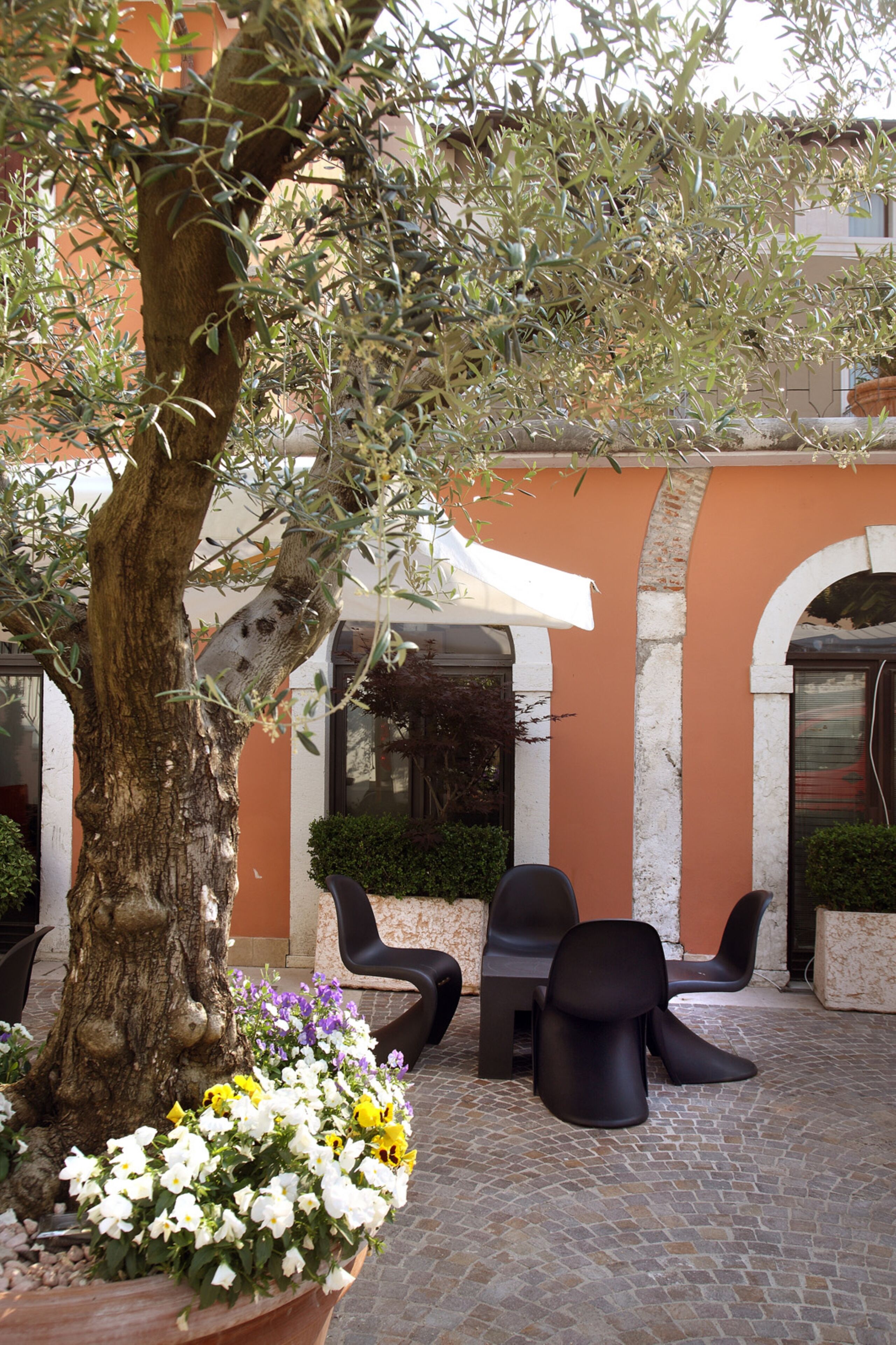 An outdoor seating area with a tree and flower pots, surrounded by a building with arches and a tiled floor.