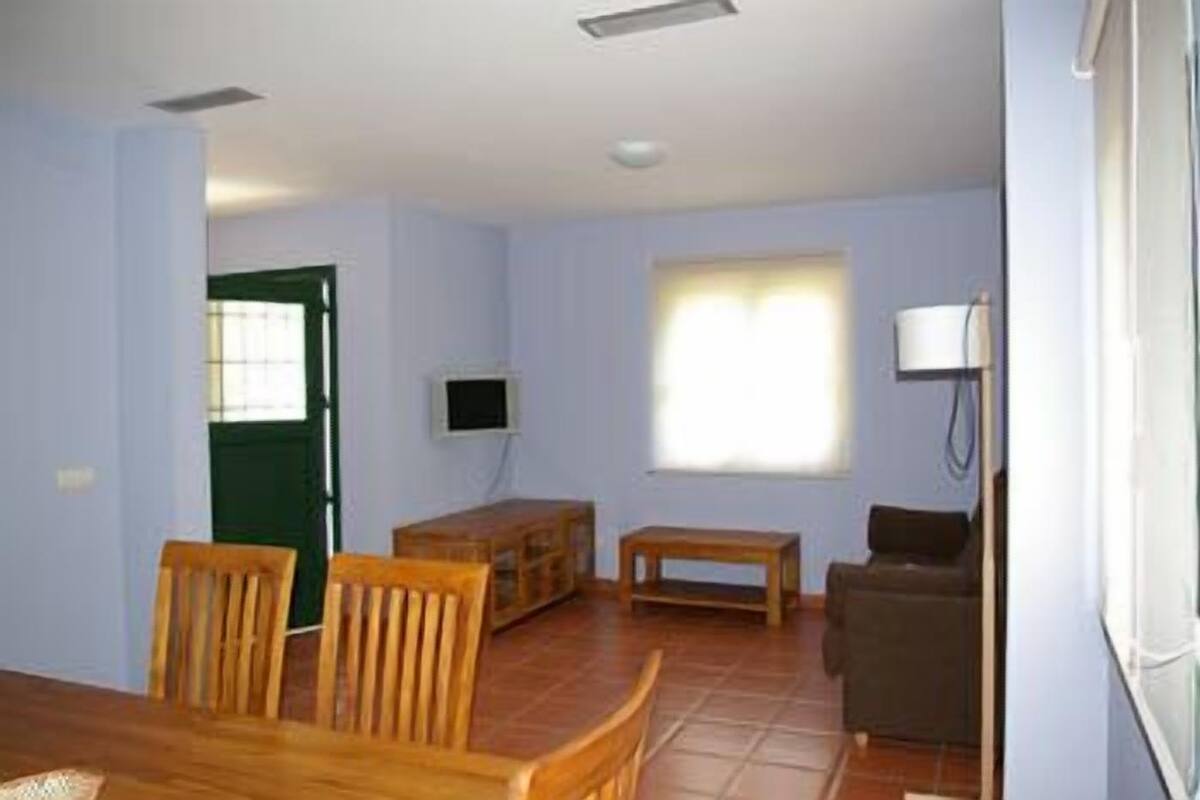 Ein Zimmer mit Esstisch und Stühlen, einer kleinen Holzbank, einem an der Wand montierten Fernseher und einem fenster mit Vorhängen.