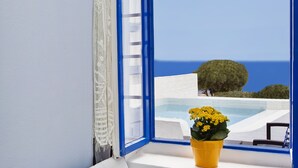Un divano bianco con cuscini verdi e multicolore, un vaso a forma di fiore giallo e vista sul mare attraverso una finestra con cornice blu.