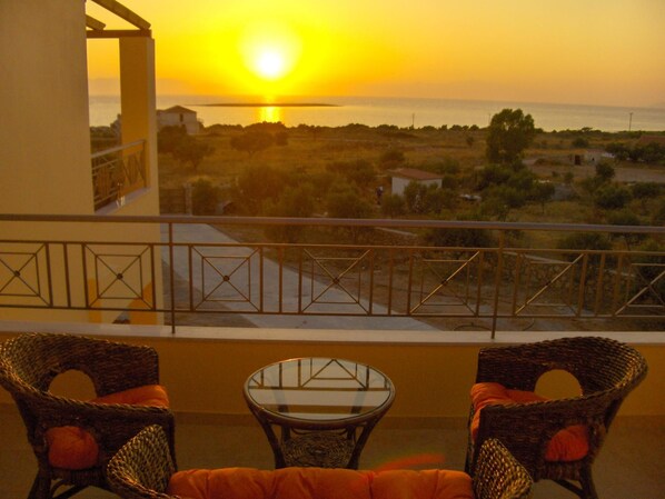 A balcony with wicker chairs and a glass table, overlooking a coastal landscape with a sunset.