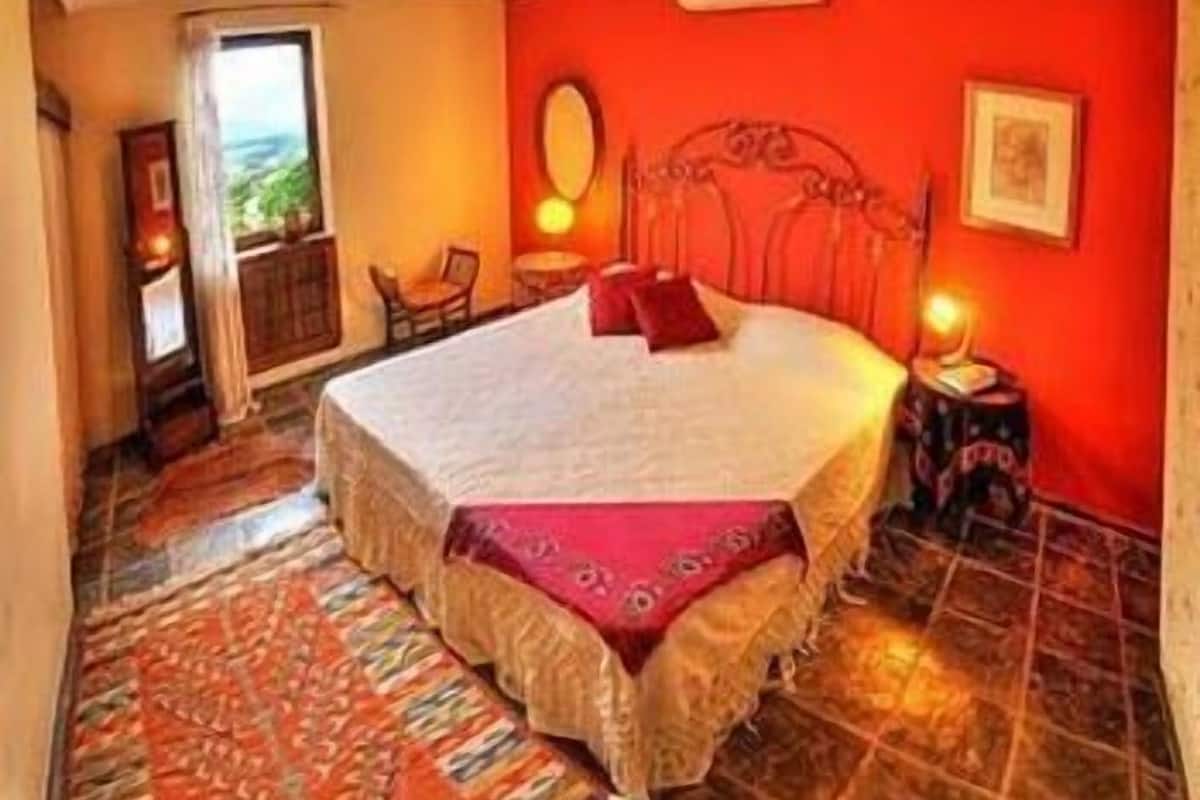 Quarto com uma cama grande, parede vermelha, janela e uma mesinha com abajur.