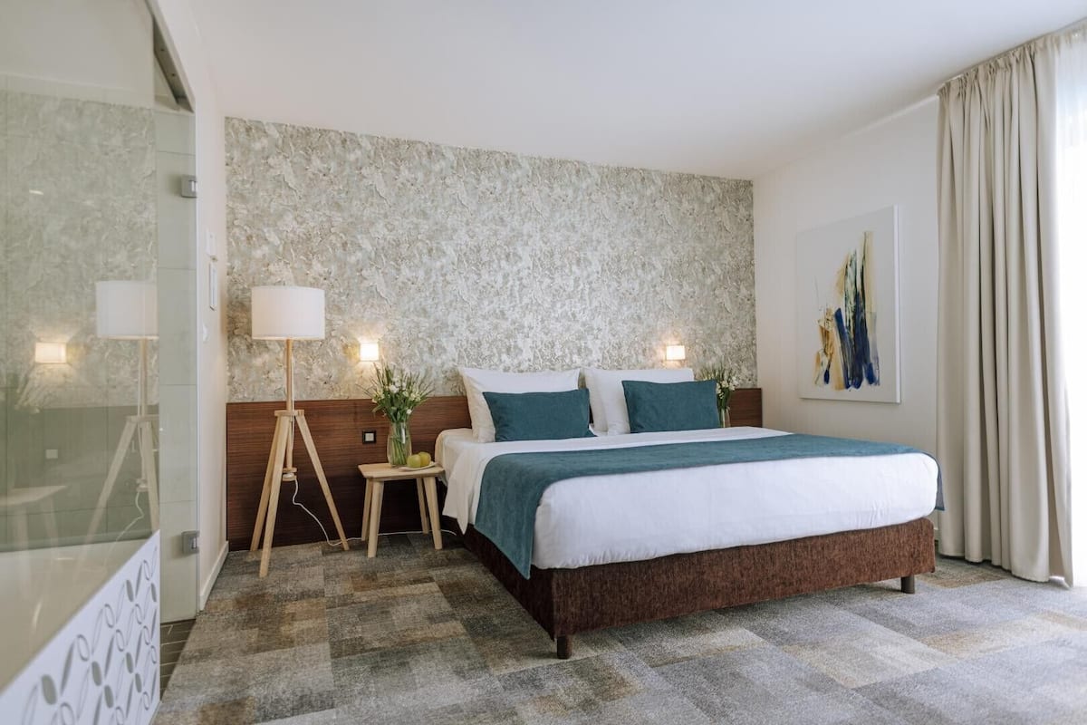 A hotel room with a large bed, a nightstand with a lamp, a small table with a vase, and a textured wall.