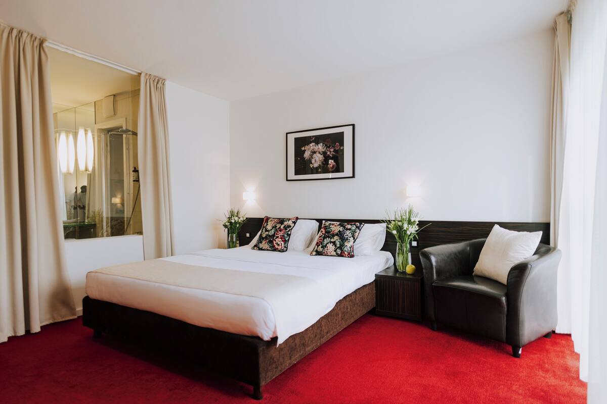 A hotel room with a large bed, a black leather armchair, a bedside table with a vase, and a framed picture on the wall.