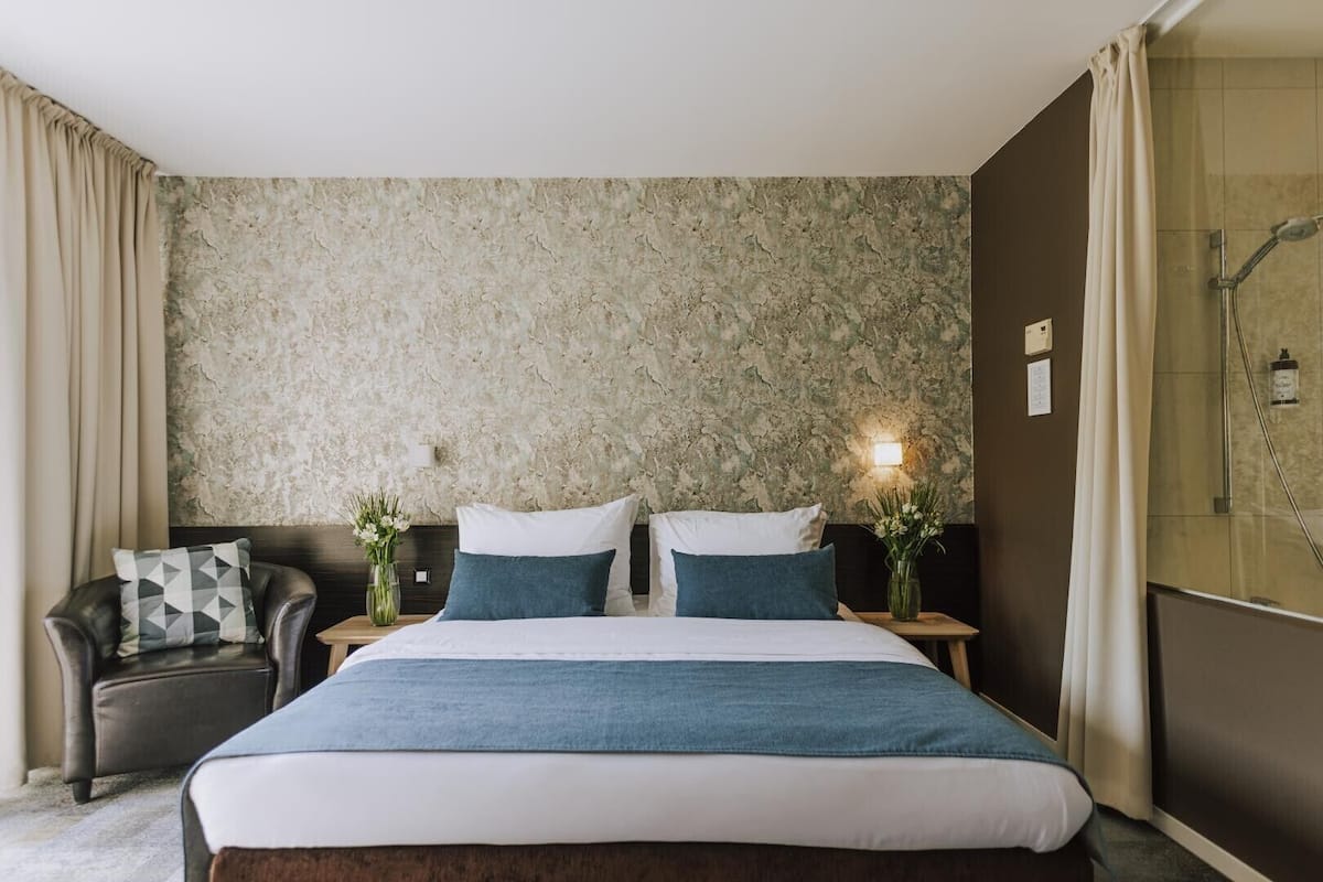 A hotel room with a large bed, two bedside tables with lamps, a chair, and a shower area.