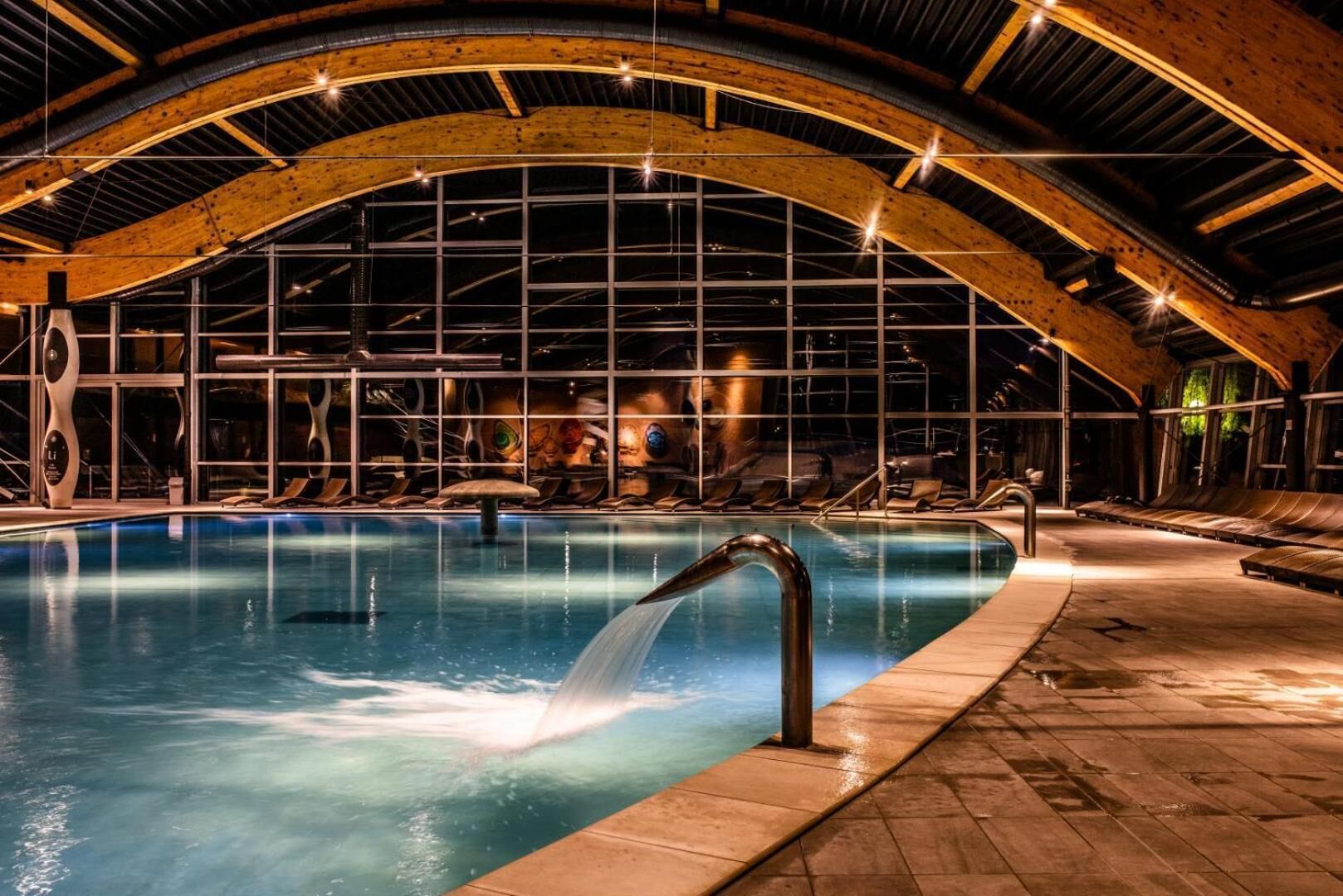 Una piscina interior con caída de agua, rodeada de sillas de descanso y una estructura de techo de madera.