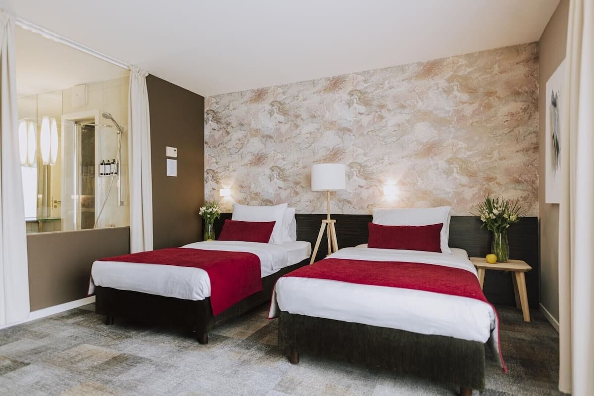 A hotel room with two beds, a marble-patterned wall, a glass shower enclosure, and a bedside table with a lamp.