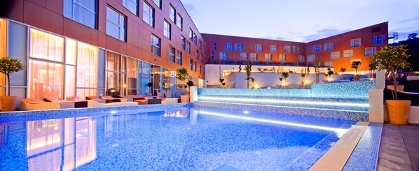 A modern hotel with a large outdoor swimming pool, lounge area with seating, and a well-lit building facade.