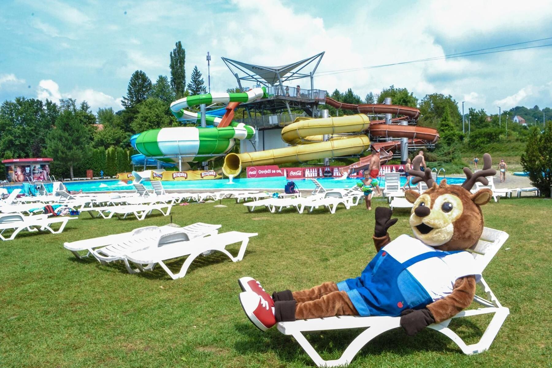 Un personaje promocional descansando en una silla de salón en un parque, con un parque acuático al fondo.