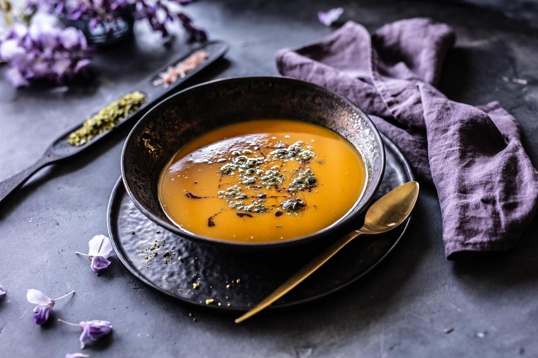 A bowl of soup with a spoon on a dark plate, surrounded by purple flowers and a cloth.