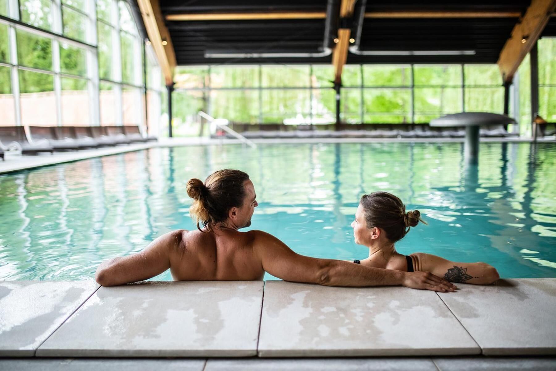 Una pareja se relaja junto a una piscina interior con vistas a la vegetación a través de amplios ventanales.