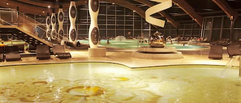 Un área de piscina interior moderna con esculturas únicas, una fuente central y una zona de estar con tumbonas.