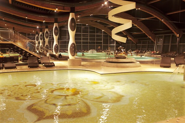 Un área de piscina interior moderna con esculturas únicas, una fuente central y una zona de estar con tumbonas.