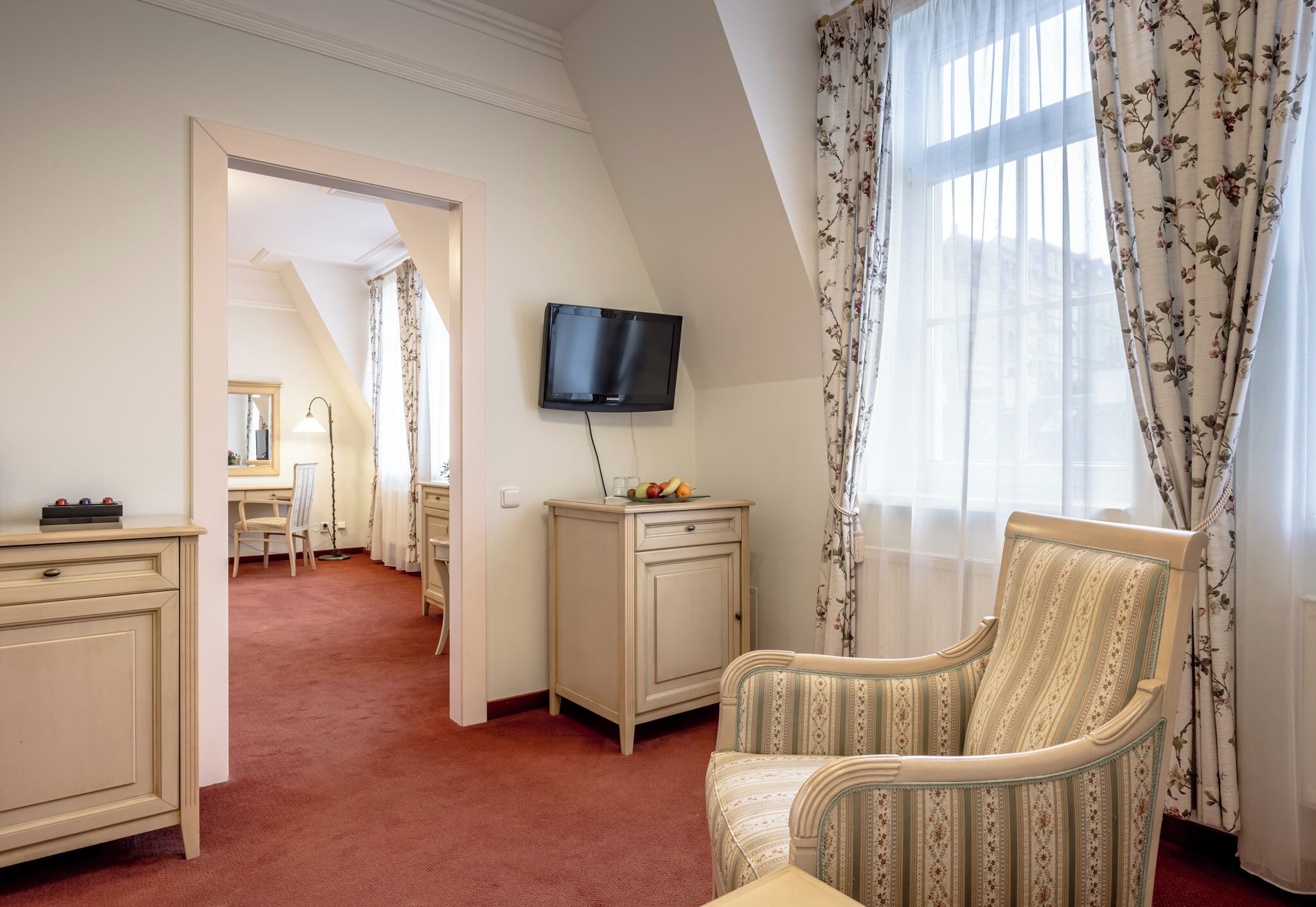 Hotellihuone, jossa on seinälle asennettu televisio, työpöytä tuolilla, kaappi ja suuri ikkuna, jossa on verhot.