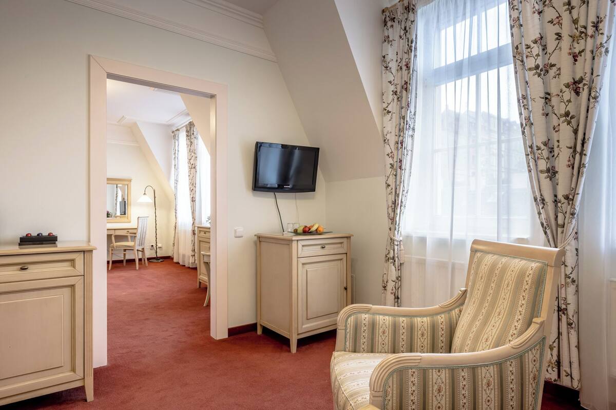 A hotel room with a TV mounted on the wall, a desk with a chair, a cabinet, and a large window with curtains.