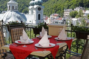 Une table dressée pour le dîner, avec une nappe rouge, des serviettes blanches et des couverts en argent, donnant sur un paysage urbain avec une église et des immeubles.