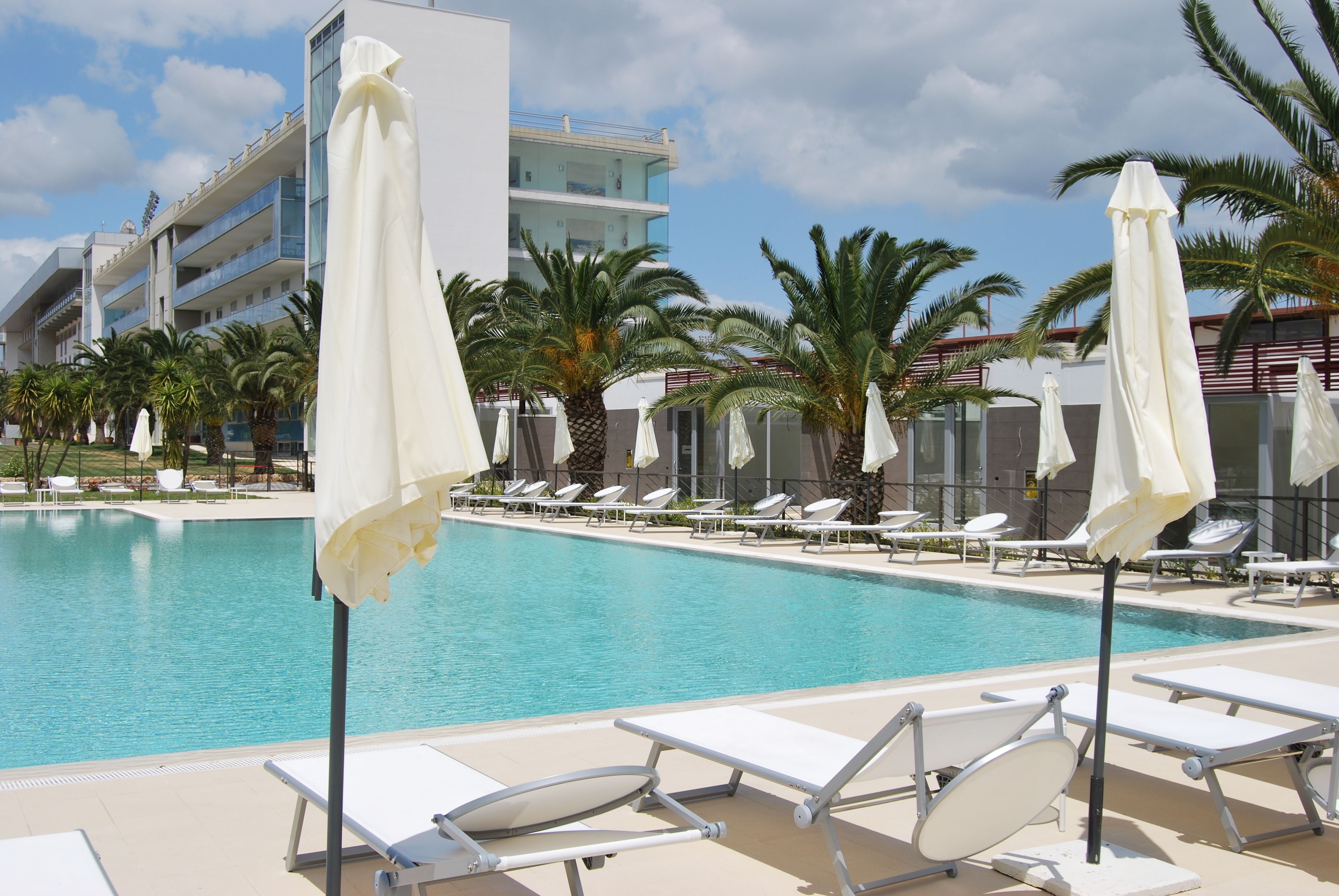 A swimming pool with lounge chairs and umbrellas, surrounded by palm trees and a modern building.