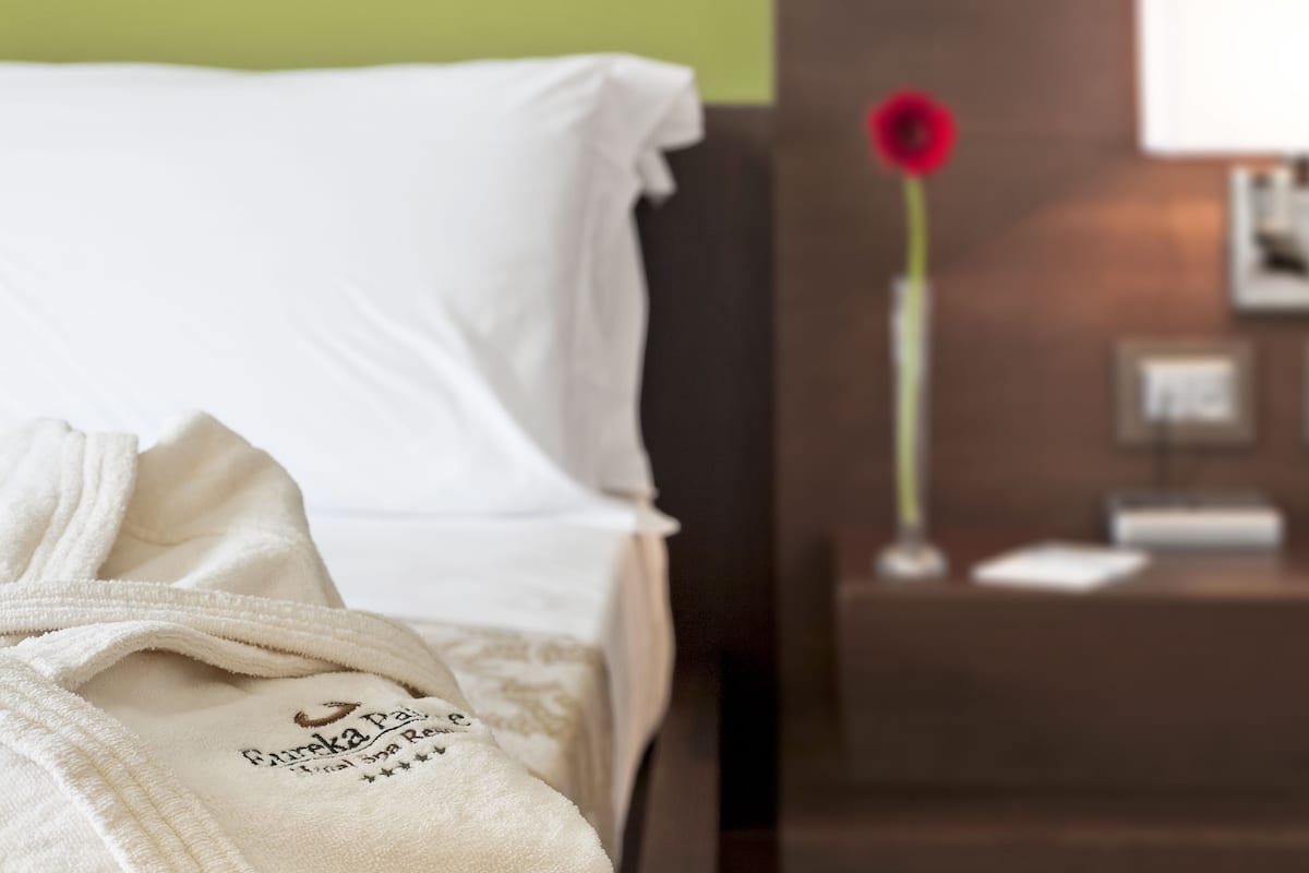 A hotel room with a neatly made bed, a bedside table with a lamp, and a red flower on the wall.
