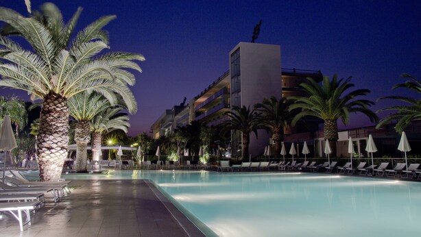 Un área de piscina nocturna con sillones de descanso y sombrillas, rodeada de palmeras y un edificio con balcones.