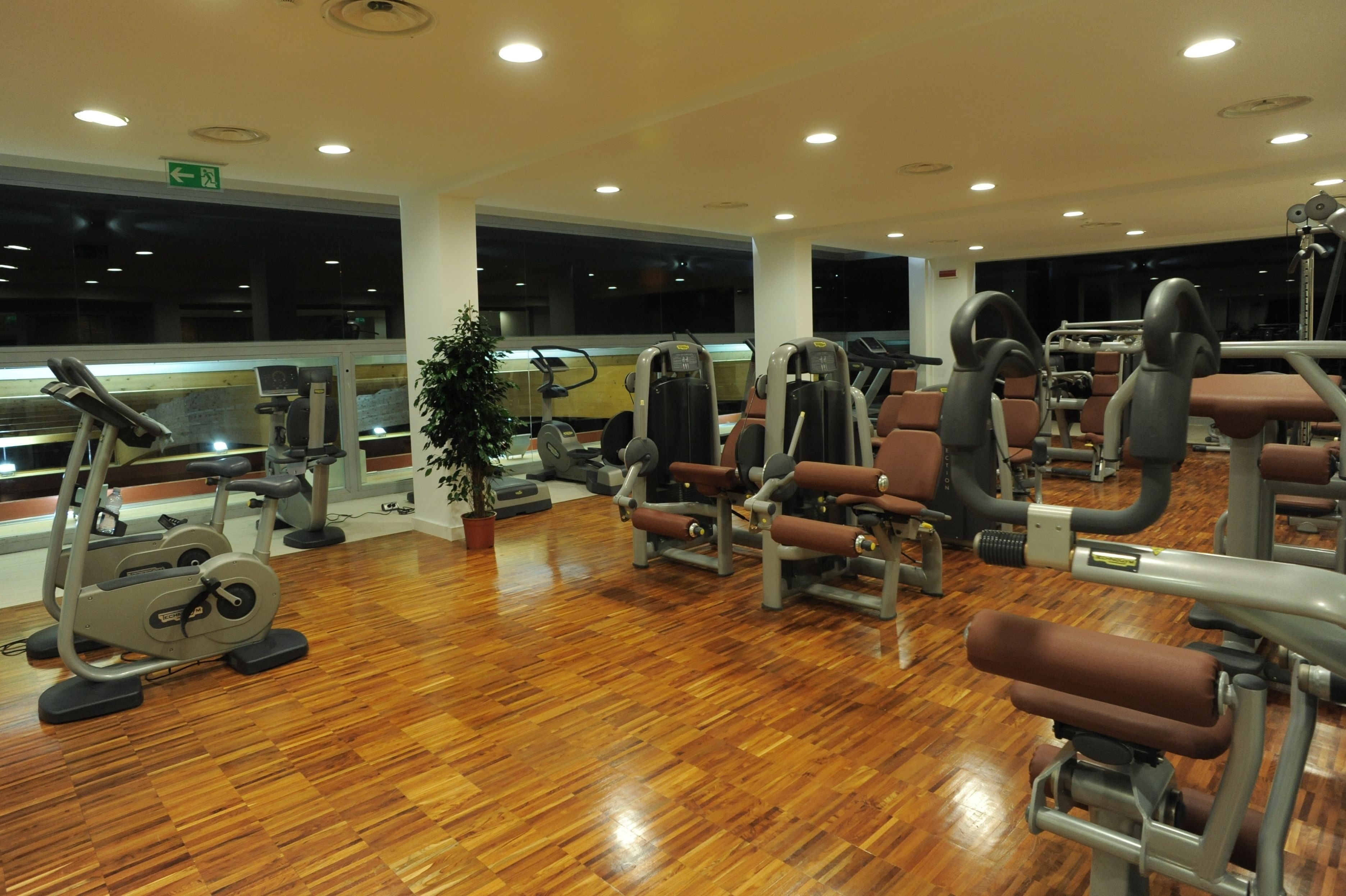 Un gimnasio con diferentes máquinas de ejercicio, suelo de madera y ventanas grandes.