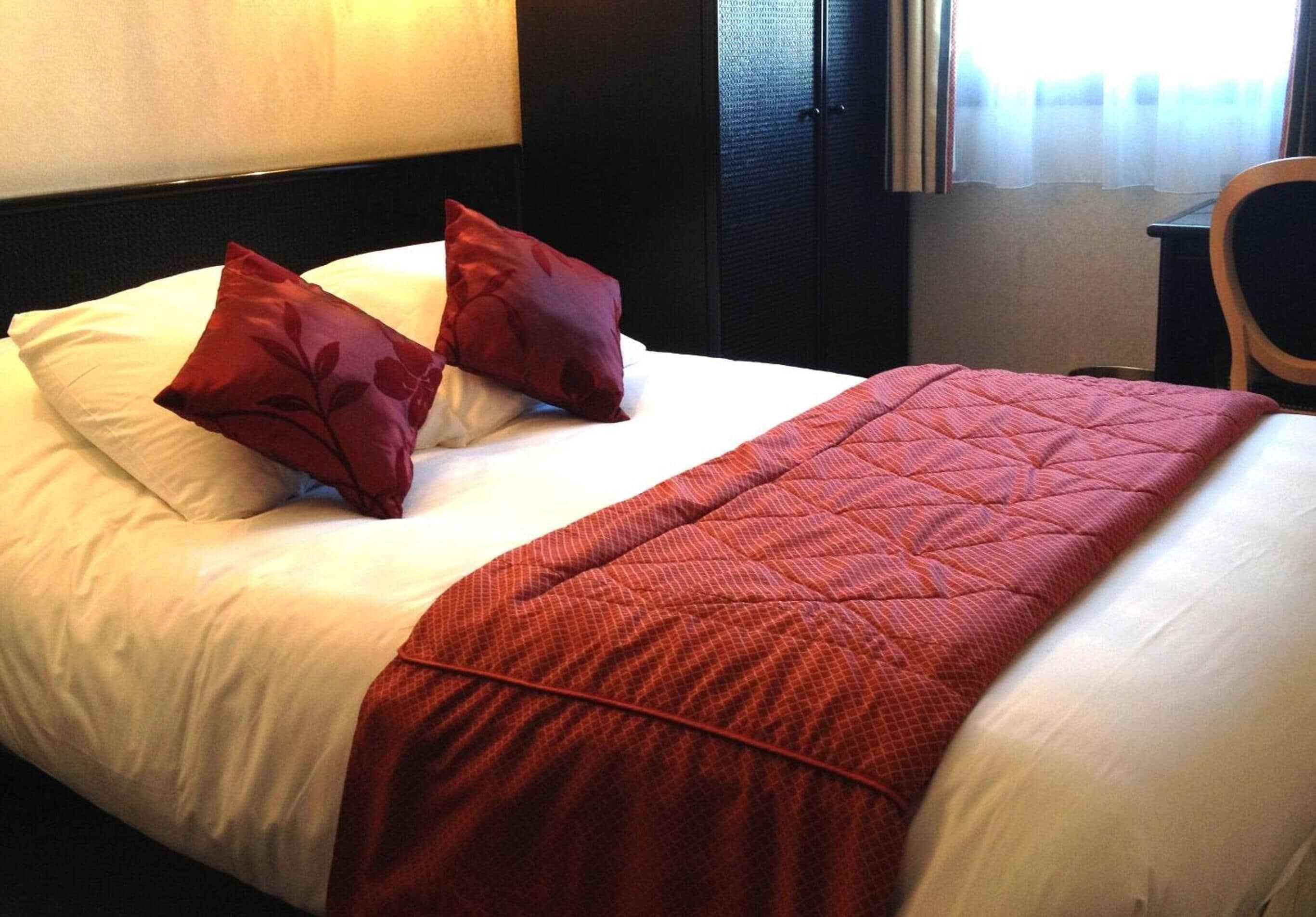 A neatly made bed with white linens and two red pillows, a red quilted blanket, and a wooden chair in a room with a dark headboard and a window with sheer curtains.