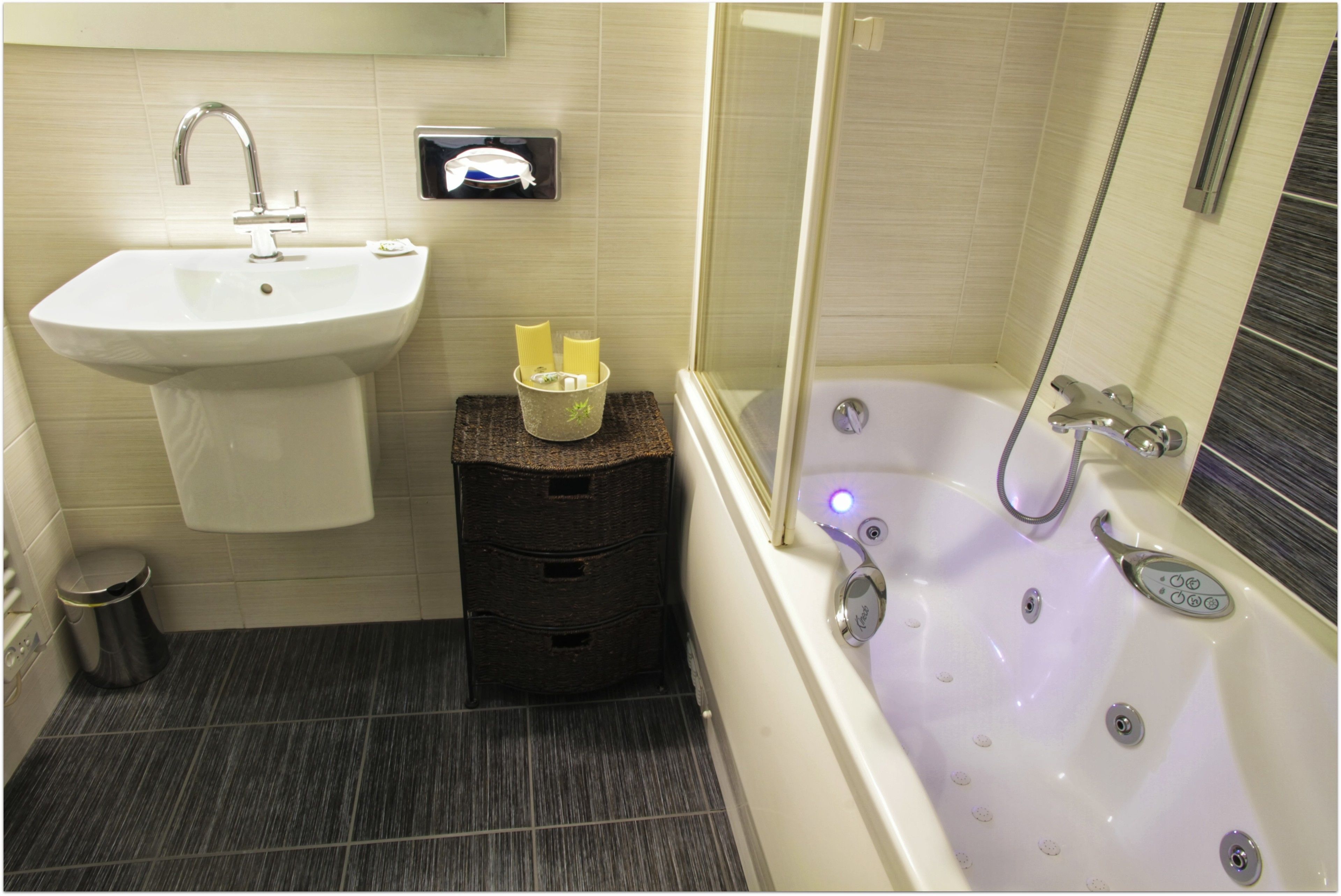 Un baño con lavamanos, un cubo de basura pequeño, un taburete de madera y una bañera con cabezal de ducha.