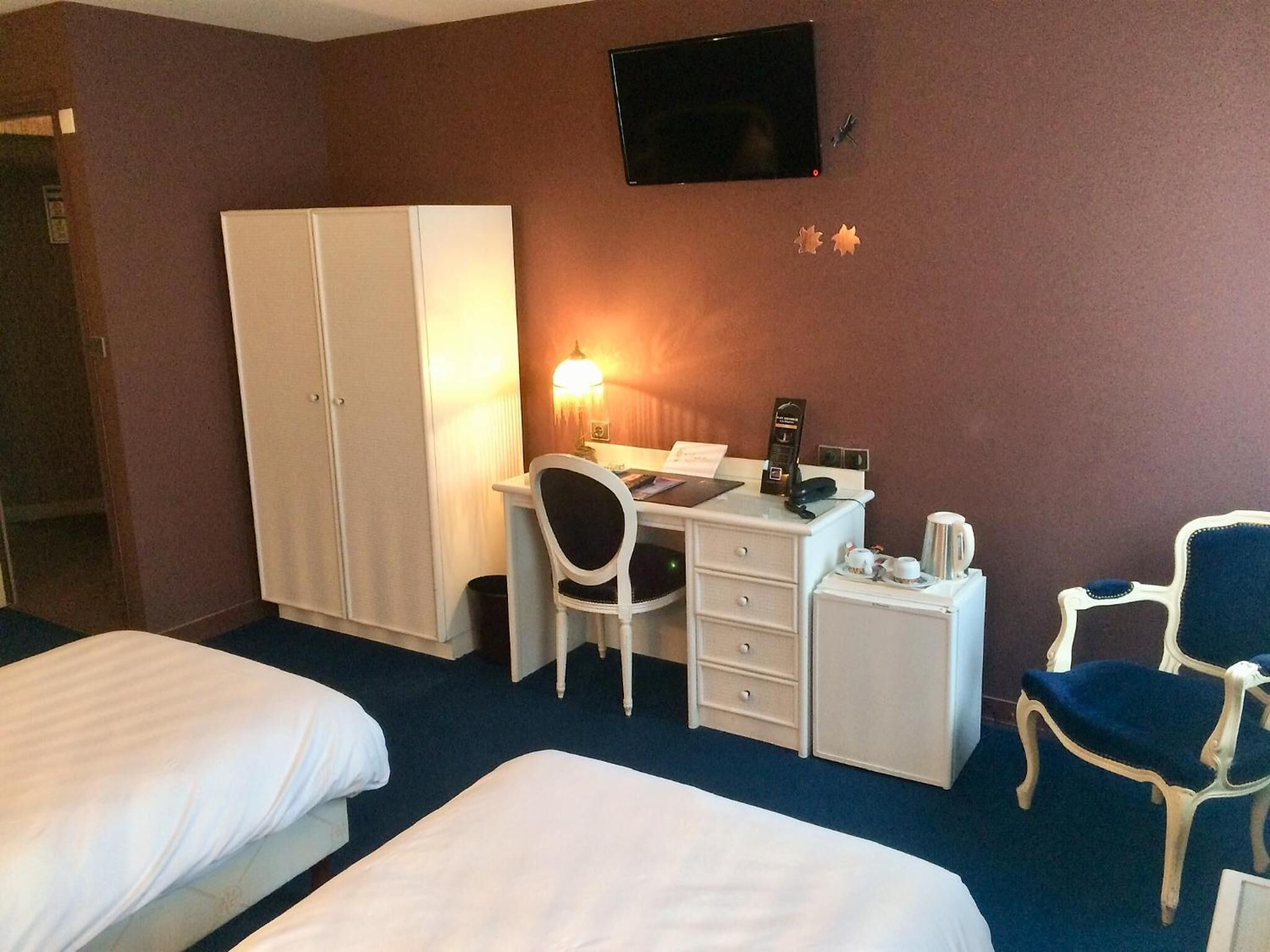 A hotel room with two beds, a desk with a chair, a wardrobe, a television, and a small table with a lamp.