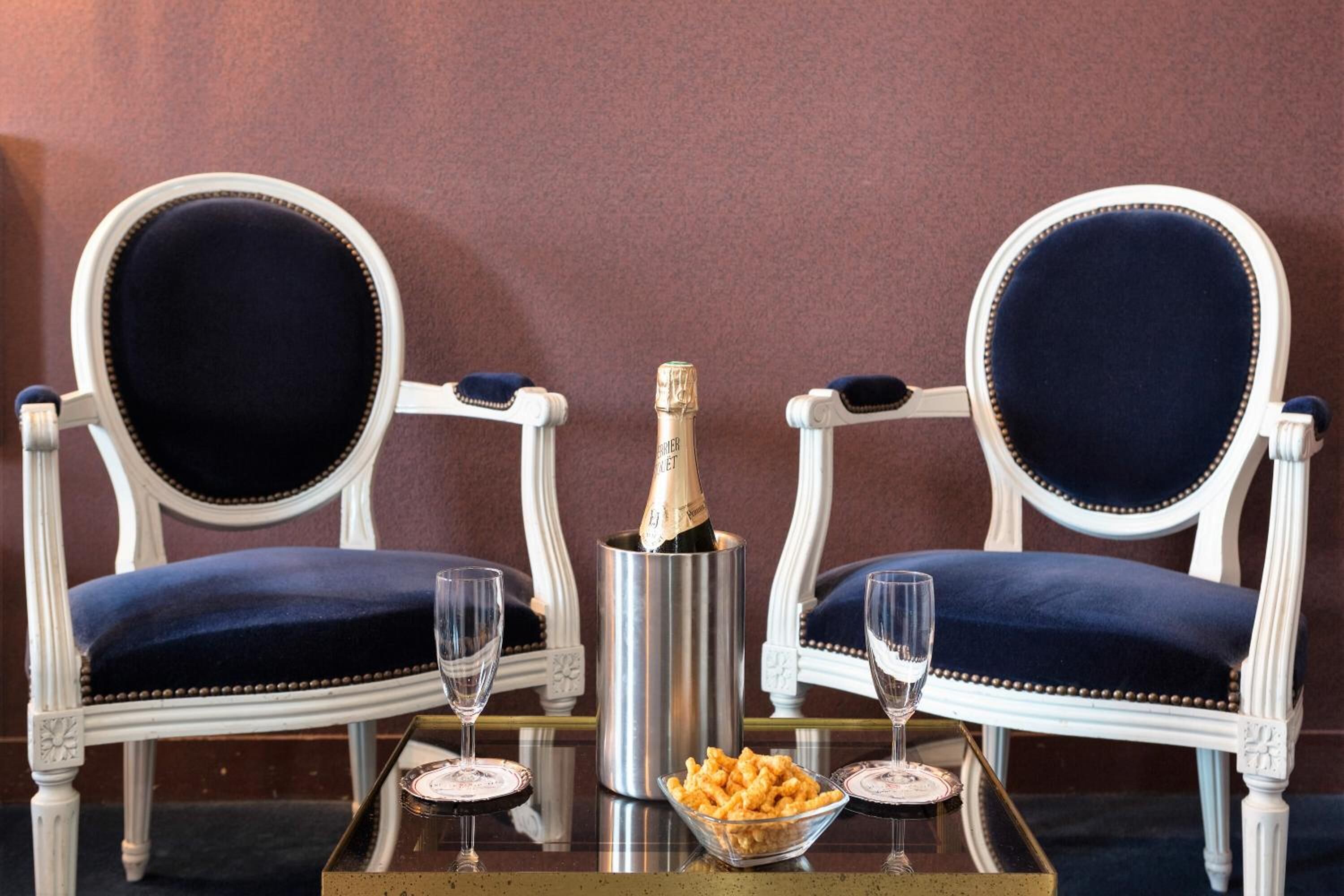 Two ornate white chairs with dark blue upholstery, a bottle of champagne, two glasses, and a bowl of snacks on a reflective table.