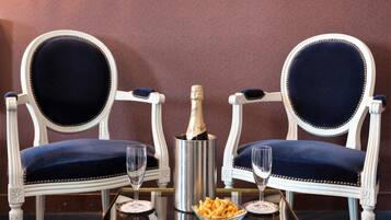 Two ornate white chairs with dark blue upholstery, a bottle of champagne, two glasses, and a bowl of snacks on a reflective table.