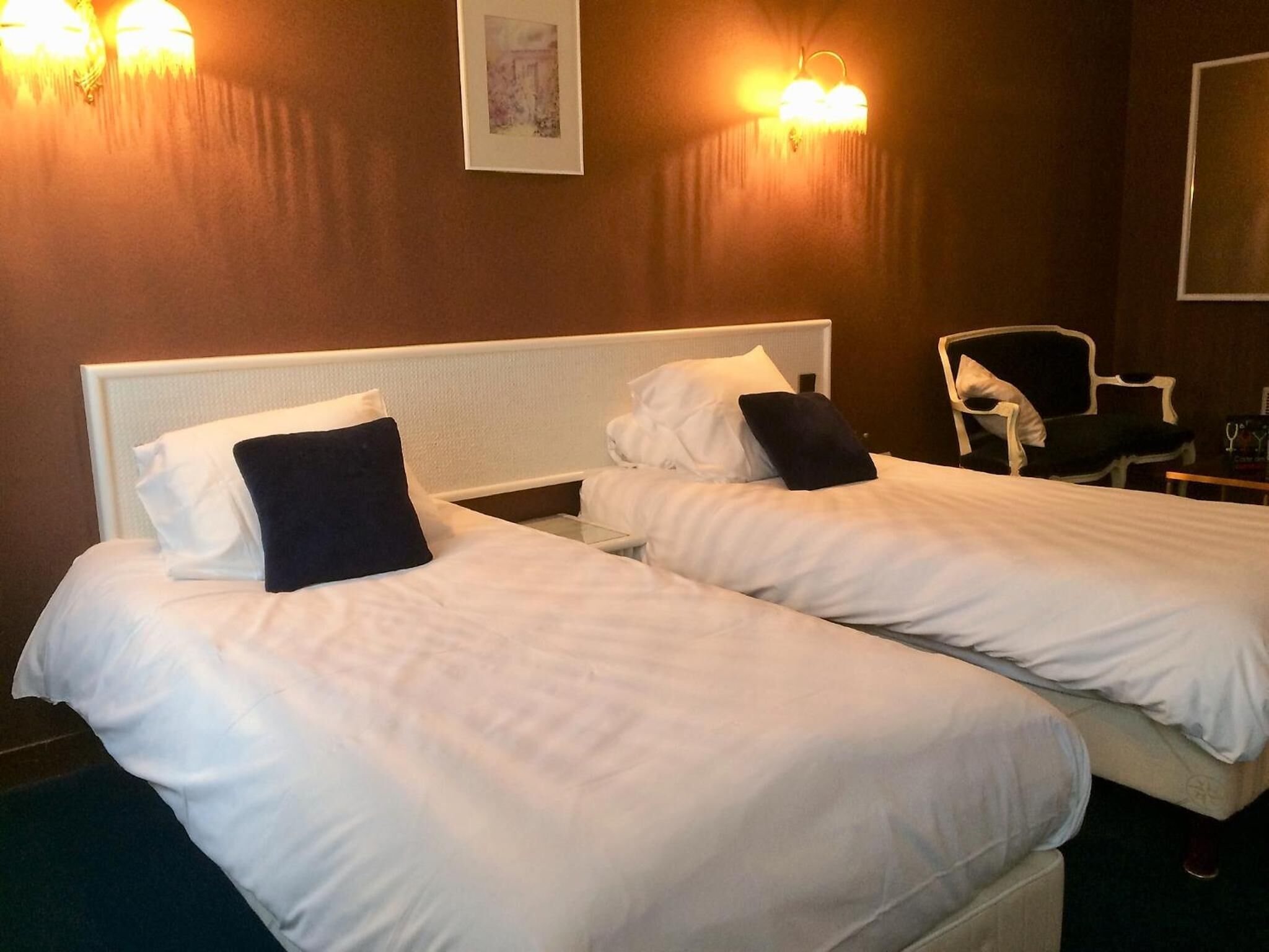 A hotel room with two single beds, each with white bedding and dark pillows. There are wall-mounted lights above the beds and a framed picture on the wall.