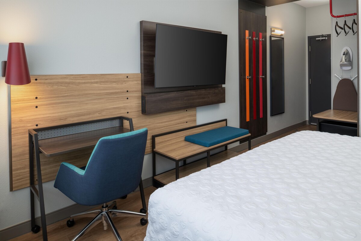 A hotel room with a bed, a desk with a chair, a TV, a mirror, and a red lamp.