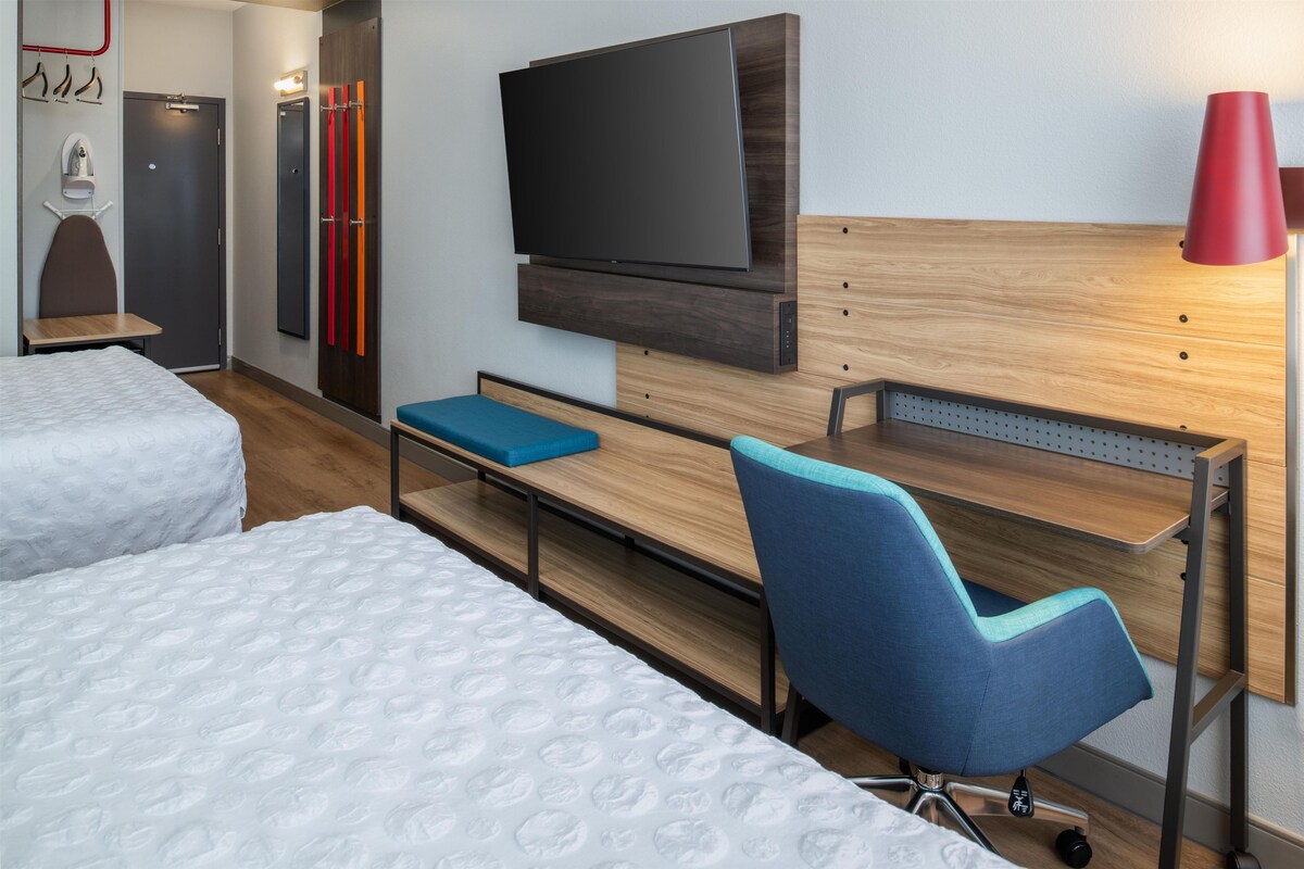 A hotel room with two beds, a TV mounted on the wall, a desk with a chair, and a red lamp.