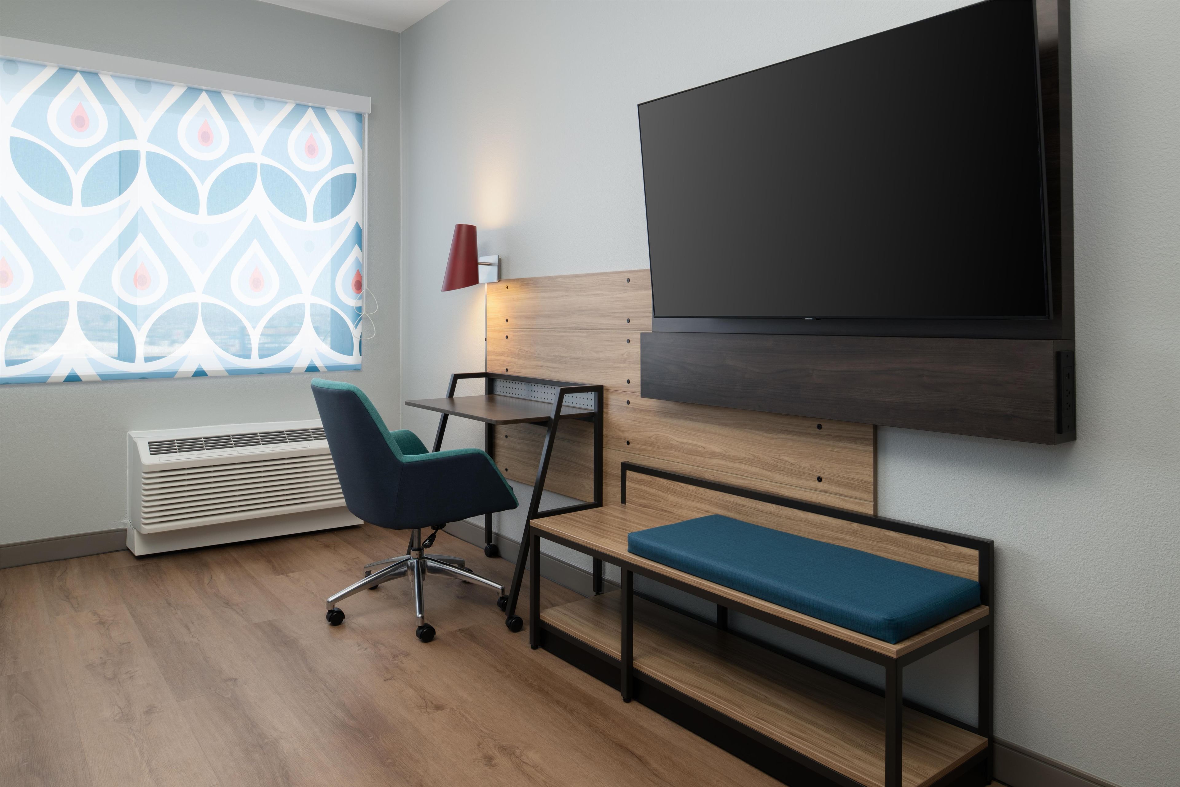 Una habitación moderna con un televisor de pantalla plana grande, un escritorio con una silla, un banco, una ventana con persianas decoradas y una lámpara roja.
