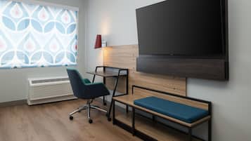 Una habitación moderna con un televisor de pantalla plana grande, un escritorio con una silla, un banco, una ventana con persianas decoradas y una lámpara roja.