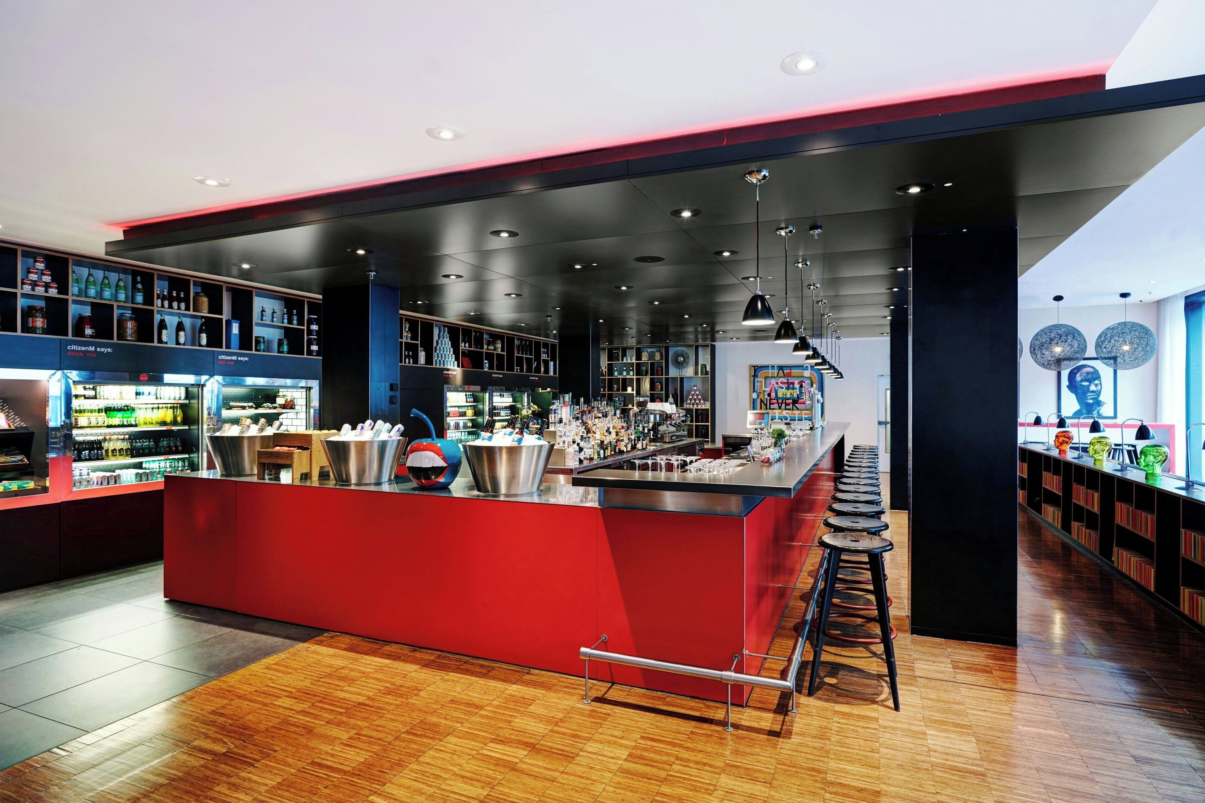 A modern bar area with a red counter, wooden flooring, and a variety of bottles and glasses on shelves.