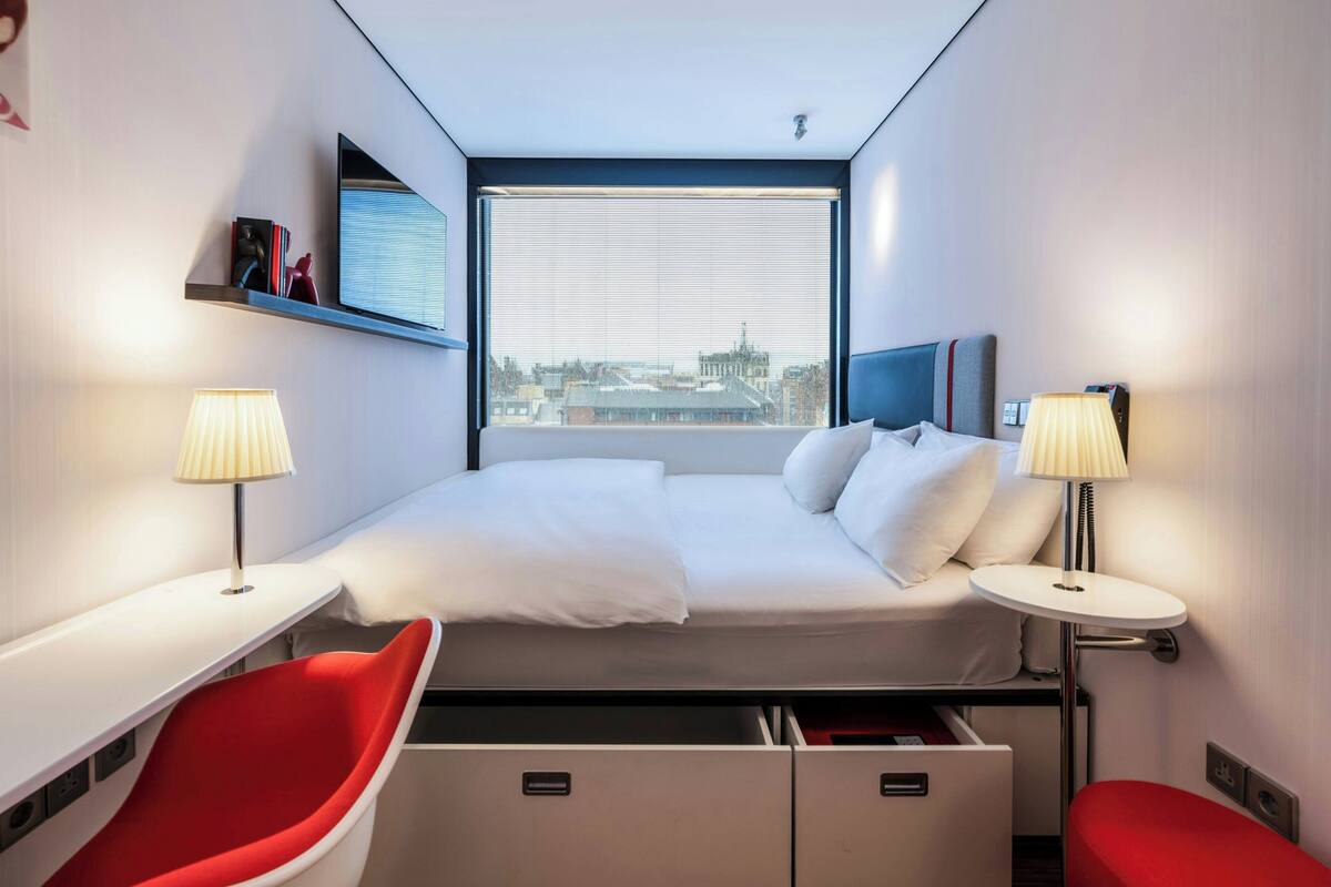 A modern hotel room with a bed, two bedside tables, a red chair, a small desk, and a window with a city view.