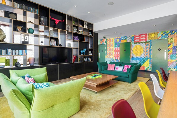 A modern living room with green sofas, a wooden coffee table, and colorful chairs. There is a bookshelf with various items and a colorful wall art piece. - citizenM Glasgow (Glasgow)