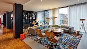 Un salon moderne avec un canapé d’angle, une table basse et un tapis coloré. On y trouve une bibliothèque remplie de divers objets et une fenêtre donnant sur des immeubles.