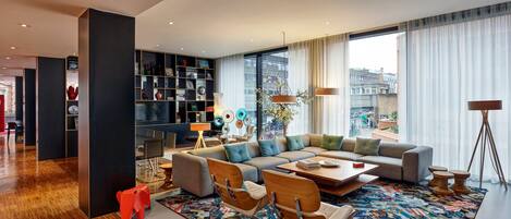 A modern living room with a sectional sofa, a coffee table, and a colorful rug. There is a bookshelf with various items and a window with a view of buildings.