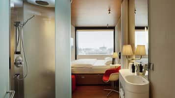 Een compacte hotelkamer met douche, een bed met uitzicht over de stad, een klein bureau en een badkamer met wastafel en spiegel.