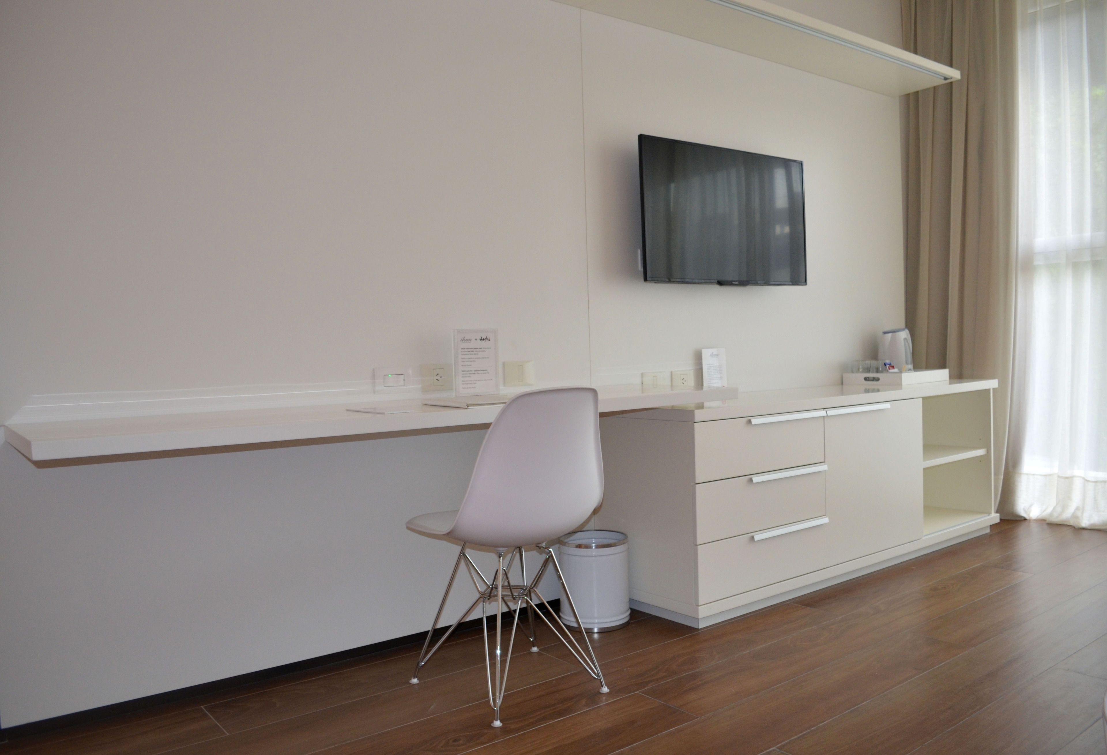 A modern hotel room with a white desk, a chair, a television mounted on the wall, and a window with curtains.