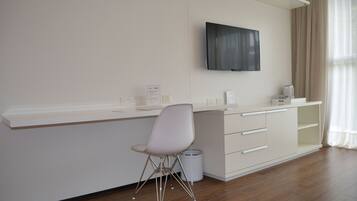 A modern hotel room with a white desk, a chair, a television mounted on the wall, and a window with curtains.