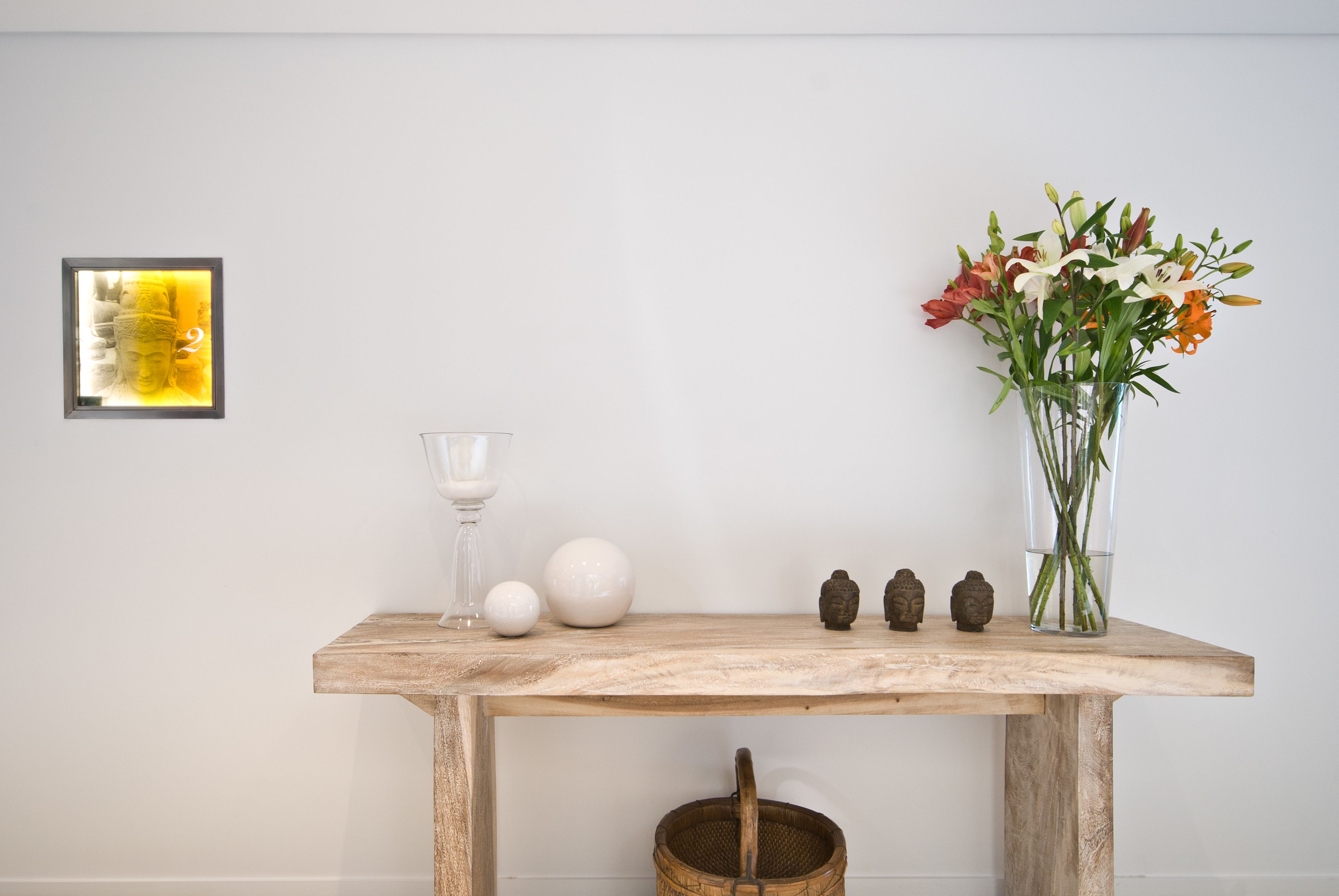 A wooden table with a glass, a white vase with flowers, a Buddha statue, and a framed picture on the wall.