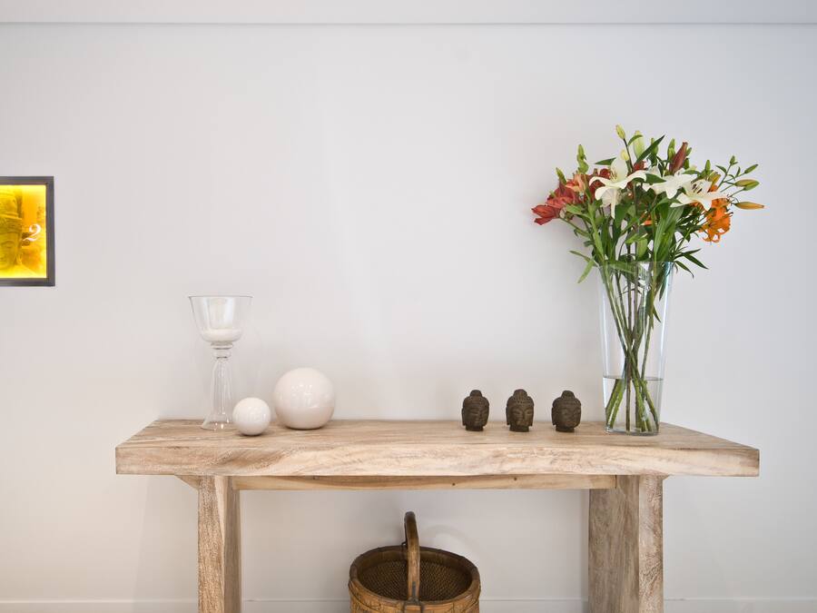 A wooden table with a glass, a white vase with flowers, a Buddha statue, and a framed picture on the wall.