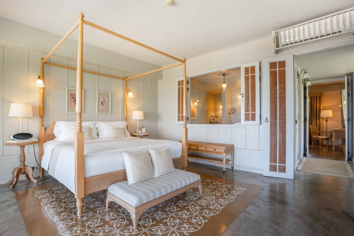 Una cama con dosel, un banco, una mesita de noche, un espejo y una puerta que da a un baño.