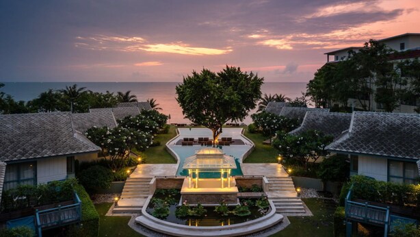 Un resort con una piscina central, rodeada de zonas de descanso y un pabellón, con vistas al océano al atardecer.
